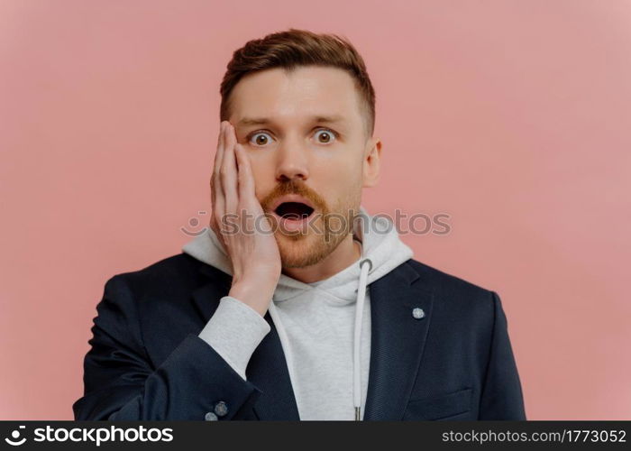 Amazed shocked handsome man with beard and red hair in jacket over hoodie dropping jaw and looking at camera with wow expression while standing isolated over pink wall. Human emotions concept. Impressed guy getting some awesome news and being in shock