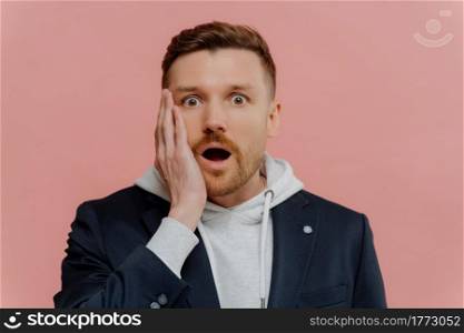 Amazed shocked handsome man with beard and red hair in jacket over hoodie dropping jaw and looking at camera with wow expression while standing isolated over pink wall. Human emotions concept. Impressed guy getting some awesome news and being in shock