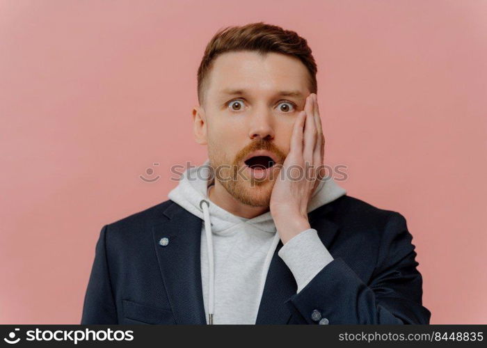 Amazed shocked handsome man with beard and red hair in jacket over hoodie dropping jaw and looking at camera with wow expression while standing isolated over pink wall. Human emotions concept. Impressed guy getting some awesome news and being in shock