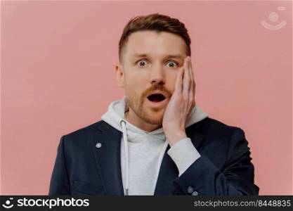 Amazed shocked handsome man with beard and red hair in jacket over hoodie dropping jaw and looking at camera with wow expression while standing isolated over pink wall. Human emotions concept. Impressed guy getting some awesome news and being in shock