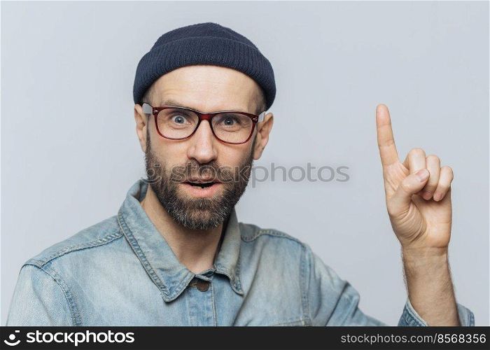 Amazed shocked man wears rectangular spectacles, looks with suprised expression, raises fore finger, has hush reaction, isolated over grey background. People, emotions and body language concept