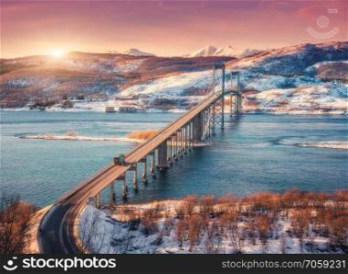 Amazing bridge during sunset in Lofoten islands, Norway. Aerial winter landscape with cars on the road, blue sea, trees, snowy mountains, colorful orange sky with clouds and sunlight. Nordic scenery