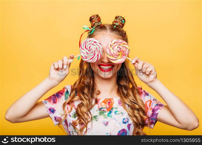 Amazing cute young girl on the yellow background holding huge sweets. Stylish girl in summer colorful dress, studio portrait. Portrait of attractive lady with two big lollypop.. Young girl on the yellow background holding sweets