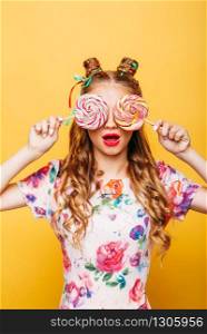 Amazing cute young girl on the yellow background holding huge sweets. Stylish girl in summer colorful dress, studio portrait. Portrait of attractive lady with two big lollypop.. Young girl on the yellow background holding sweets