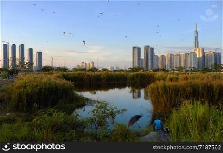 Amazing landscape of Ho Chi Minh city, Vietnam, many kites flying on sunset sky, high rise building reflect on water, reeds flowers and man sit for fishing at foreground make wonderful scene