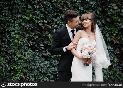 Amazing smiling wedding couple. Pretty bride and stylish groom. groom and bride posing in front wall with green ivy.. Amazing smiling wedding couple. Pretty bride and stylish groom. groom and bride posing in front wall with green ivy
