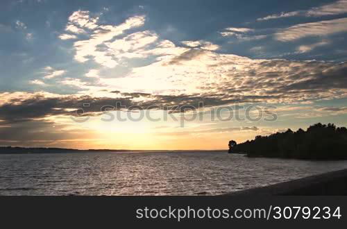 Amazing sunset above the sea and cloudscape in rays of setting sun on a windy summer day. Coastal scenery with waves breaking on the dike and forest in afterglow.