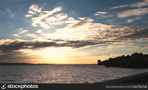 Amazing sunset above the sea and cloudscape in rays of setting sun on a windy summer day. Coastal scenery with waves breaking on the dike and forest in afterglow.