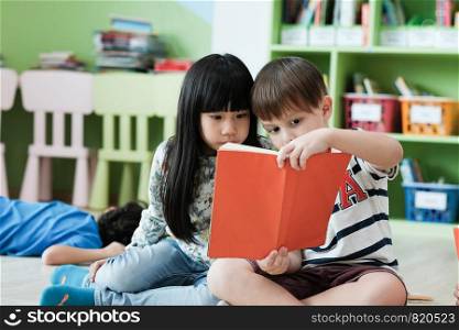 American boy and asian girl are reading together with happiness in their kindergarten classroom, kid education and diversity concept