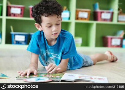 American boy lying and reading book in kindergarten classroom, kid education concept