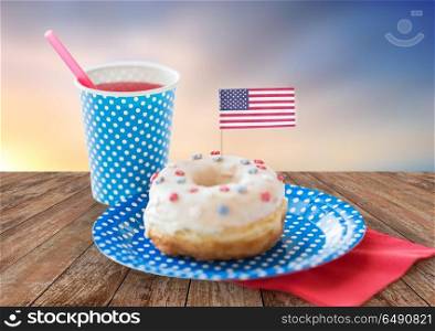 american independence day, celebration, patriotism and holiday concept - close up of glazed sweet donut decorated with flag and juice in disposable cup at 4th july party over evening sky background. donut with american flag decoration and drink. donut with american flag decoration and drink