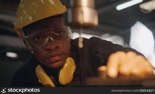 American industrial black young worker man with yellow helmet and ear protection manual rotating on drill machine In order to penetrate workpiece, Engineer at work in industry factory.