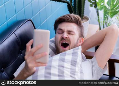 american man watching media content in a smart phone and yawn on a couch at home.