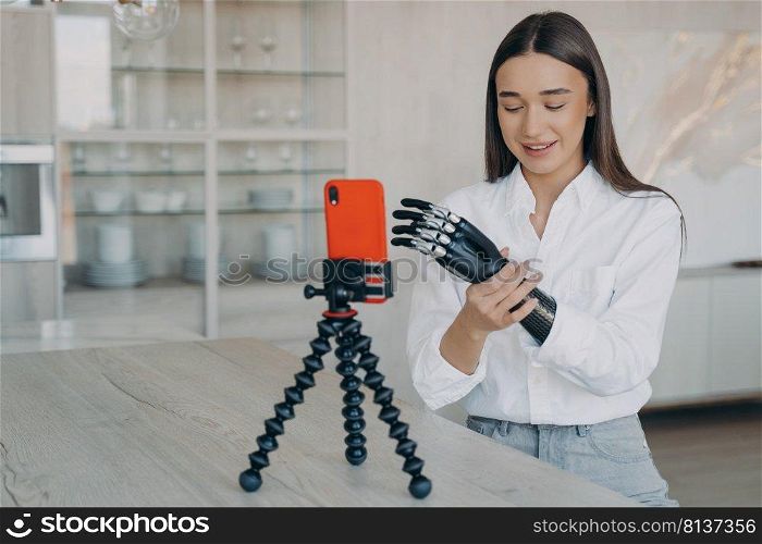 Amputee is taking video how to set a cyber arm. Attractive handicapped girl is blogger. European woman is filming with telephone on tripod. Modern bionic prosthesis using, tutorial filming.. Amputee is taking video how to set a cyber arm. Attractive handicapped girl is blogger.