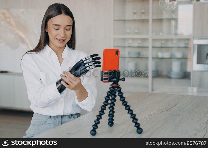 Amputee is taking video how to set a cyber arm. Attractive handicapped girl is blogger. European woman is filming with telephone on tripod. Modern bionic prosthesis using, tutorial filming.. Amputee is taking video how to set a cyber arm. Attractive handicapped girl is blogger.