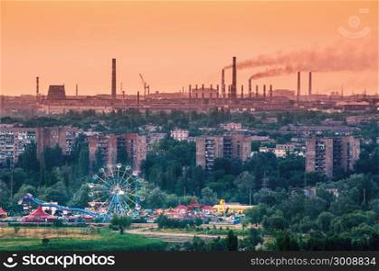 Amusement park with Ferris wheel and buildings on the background of metallurgy plant with smog at sunset. Ecology problems, atmospheric pollutants. Pipes with smoke. Heavy industry factory. Steel mill