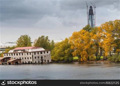 An abandoned wooden building in the water against the background of trees and a modern high-rise building.