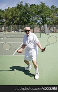 An active senior man on a tennis court with two rackets looking for someone to play.