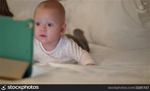 An adorable baby girl is lying on tummy on a big white bed. Her bright big grey eyes are looking at tablet that is located right in front of her. The girl is wearing a white polka dot shirt and comfy grey pants
