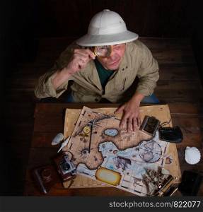An adult explorer in a cork helmet over an old wooden table paves a route using old maps. Cork Helmet Explorer
