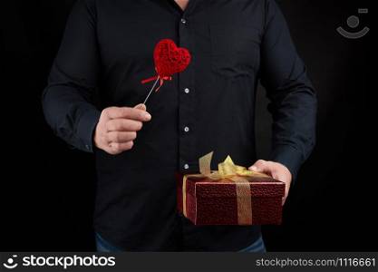 an adult man in a black shirt holds in his hand a red cardboard box tied with a golden bow and a red heart, holiday greetings concept, surprise