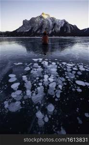 An adventurer sits in front of an abstract array of the frozen ice bubbles of Abraham Lake as the sun sets on the day.