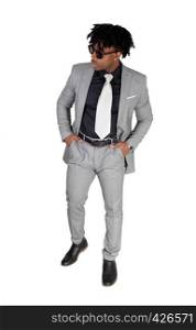 An African American young man standing in a gray suit, wearing sunglasses with his hands in his pocket, isolated for white background