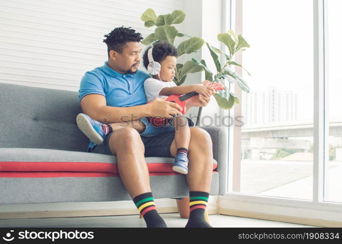 An african black father teaching and playing ukulele with his little son in living room at home. Education and Family concept.