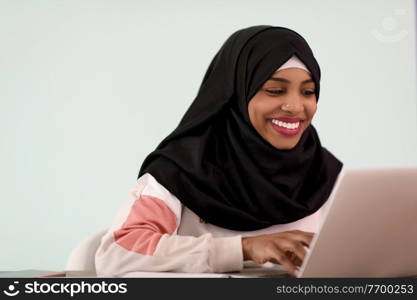 an afro muslim  woman  wearing a hijab sits smiling in her home office and uses a laptop
