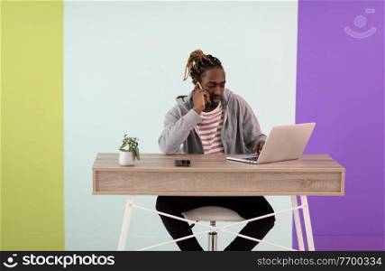 an afro young man sits in his home office during a pandemic and uses the phone to talk
