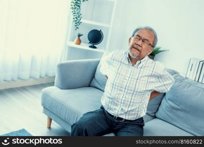 An agonizing senior man in need of assistance while sitting on his sofa at home, suffering from back pain. Senior care, nursing home for pensioners, deteriorating health of old age.. An agonizing senior man is holding his back due to back pain.
