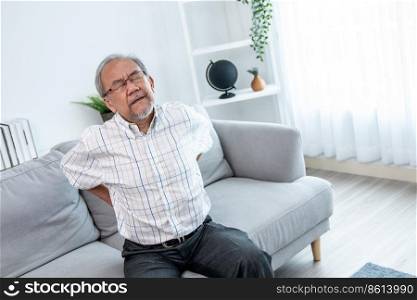 An agonizing senior man in need of assistance while sitting on his sofa at home, suffering from back pain. Senior care, nursing home for pensioners, deteriorating health of old age.. An agonizing senior man is holding his back due to back pain.