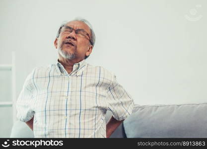 An agonizing senior man in need of assistance while sitting on his sofa at home, suffering from back pain. Senior care, nursing home for pensioners, deteriorating health of old age.. An agonizing senior man is holding his back due to back pain.