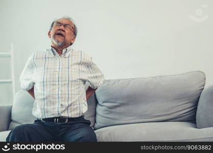 An agonizing senior man in need of assistance while sitting on his sofa at home, suffering from back pain. Senior care, nursing home for pensioners, deteriorating health of old age.. An agonizing senior man is holding his back due to back pain.