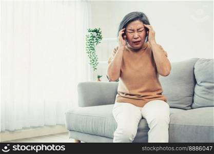 An agonizing senior woman with a headache, compress her temple with both hands at her contented living room. Senior health, nursing home, caretaker service.. An agonizing senior woman with a headache in her contented environment.