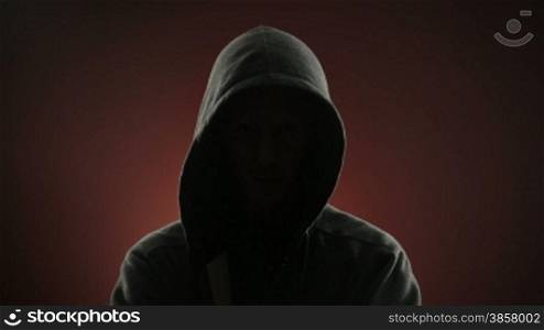 An anonymous, threatening thug wearing a hood stares at the camera in front of a red background. Off-centered version also available.