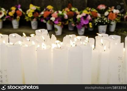 An array of white candles burning on an altar in a Buddhist temple with flower offerings arranged in the background.
