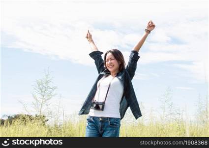 An Asian beautiful woman wearing check shirt, camera and backpack for traveling in summer time. She feels happy and refresh with background of blue sky and green field