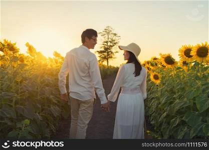 An Asian couple enjoying, relaxing, walking, holding hands each other in sunflower field on road during travel holidays vacation trip outdoors at natural garden park at sunset in Lopburi, Thailand