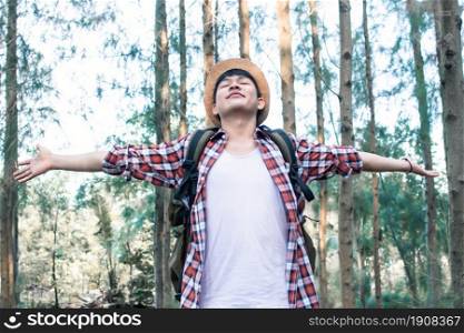 An Asian handsome man wearing hat, check shirt, camera and backpack for traveling and camping in forest for summer time. He feels happy and refresh.