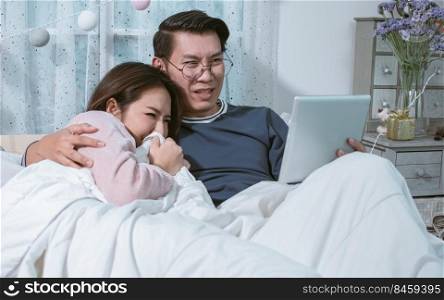 An asian lovely couple watching horror or scary movie in tablet together while laying down on bed at home. Lifestyle and Technology Concept.