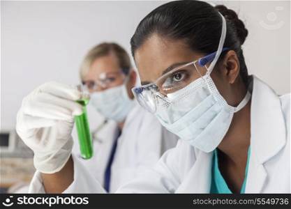 An Asian medical or scientific researcher or doctor using looking at a test tube of green liquid in a laboratory with her female colleague out of focus behind her.