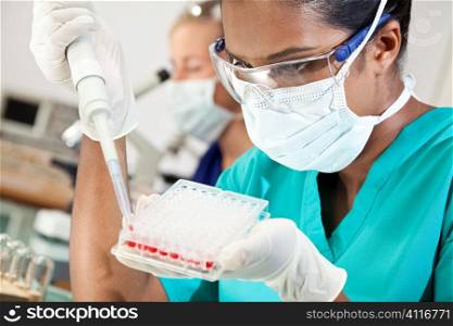 An Asian medical or scientific researcher or doctor working with a pipette, blood samples and a well tray in a laboratory with her female colleague out of focus behind her.