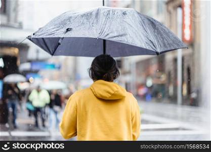 An Asian middle aged man with black grey long hair in yellow jumper hoodie holding black umbrella waiting for traffic light in city, during raining day.