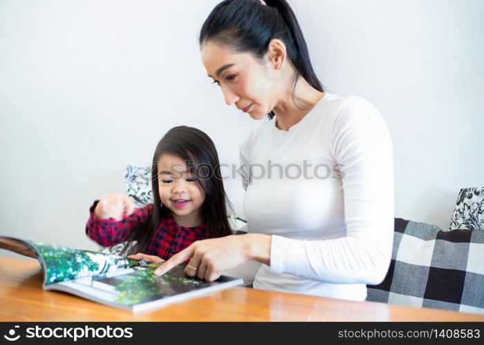 An Asian mother is teaching her daughter to read a book during the semester break on the living table and having cold milk on the table at home. Educational concepts and activities of the family