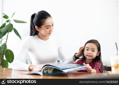An Asian mother is teaching her daughter to read a book during the semester break on the living table and having cold milk on the table at home. Educational concepts and activities of the family