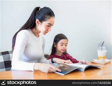 An Asian mother is teaching her daughter to read a book during the semester break on the living table and having cold milk on the table at home. Educational concepts and activities of the family