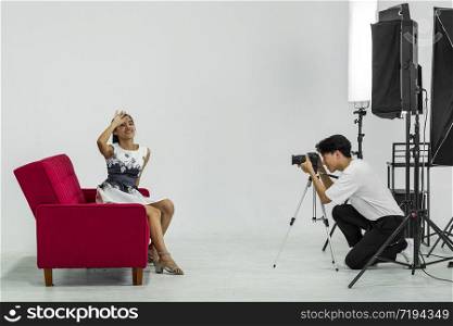 An Asian photographer is photographing a model posing on a red sofa for advertising in a magazine.