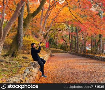 An Asian tourist man, a photographer, using a camera to take photos at red maple leaves or fall foliage in colorful trees in autumn season in travel trip and holidays vacation concept. Kyoto, Japan.