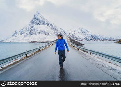An Asian tourist man walking on road and traveling in Lofoten islands, Norway, Europe. White snowy mountain hills, nature landscape in winter. Trekking and hiking. Adventure.